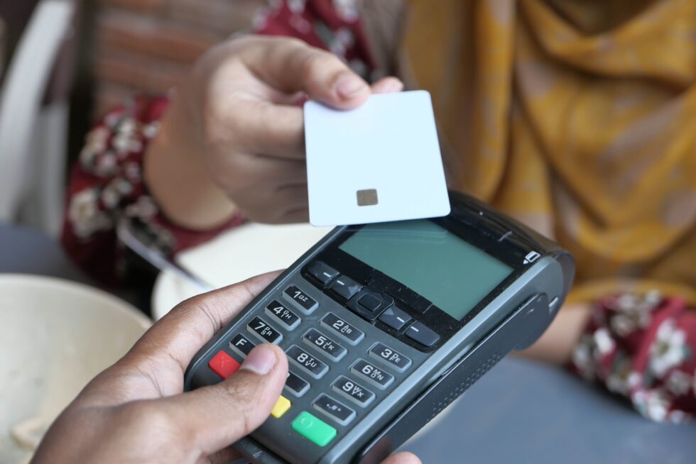 A hand holds a credit card above a payment terminal, illustrating the process of credit card processing. Optimized Payments enhances authorization strategies to reduce processing fees effectively.